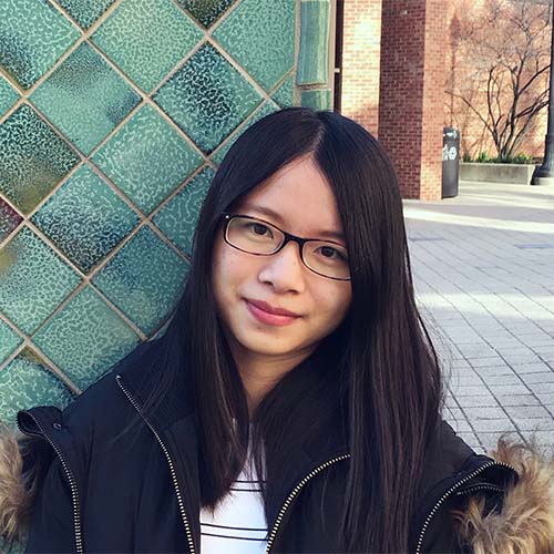 A headshot of CS assistant professor Ziyu Yao, wearing glasses, a dark winter coat with a fur-trimmed hood, stands outdoors near a green-tiled wall and brick buildings.